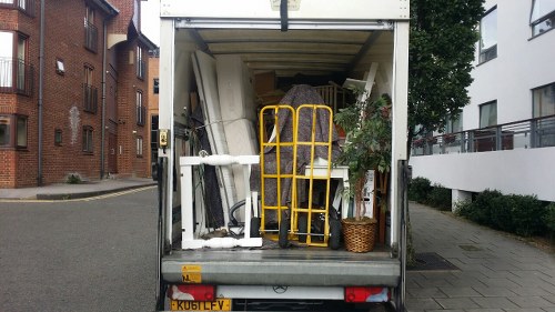 The image shows the open rear of a commercial waste removal vehicle parked on a residential street in Highgate, with a variety of discarded items piled inside. Visible objects include white and black plastic pipes, a yellow hand truck or dolly standing upright in the centre, and a woven wicker basket positioned towards the right side. The waste materials appear to be a mixture of household or business refuse, such as pipe segments, metal parts, and possibly bits of packaging or packaging materials. The scene is set against a background of urban buildings, with brick walls on the left and modern white houses with balconies on the right, indicating a typical residential area in North London. The weather appears clear with natural daylight illuminating the scene, emphasizing the textures and colors of the waste and surroundings. As part of their rubbish collection service, Commercial Waste Highgate is depicted managing waste disposal in this local area, integrating clearly visible items with urban infrastructure.