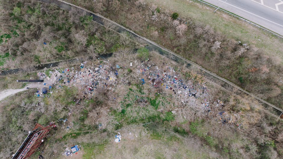 An aerial view of a large, irregularly shaped area covered with scattered debris and rubbish, including plastic bags, bottles, and miscellaneous waste, spread across a patch of land with some sparse g
